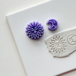 May include: Purple plastic sun and moon cookie cutters on a white surface. The sun and moon have faces.