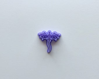 May include: A purple dragon-shaped charm with a white outline. The charm is made of a soft, flexible material.
