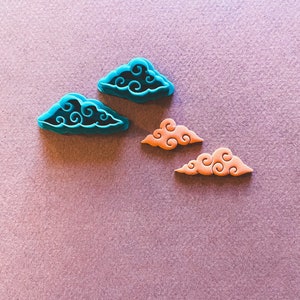 Puede incluir: Cuatro cortadores de galletas con forma de nubes con detalles en espiral. Dos son azules y dos son naranjas.