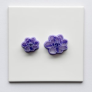 Puede incluir: Dos cortadores de galletas morados con forma de flor. Los cortadores son de plástico y tienen un diseño floral detallado. El cortador más pequeño está a la izquierda del más grande. El fondo es un cuadrado blanco.