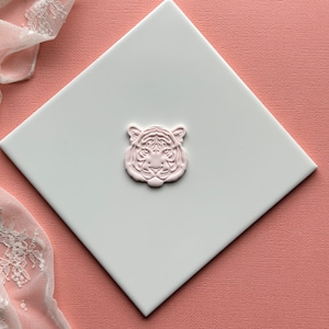 May include: A pink ceramic tiger head ornament on a white square tile. The tile is on a pink textured surface.
