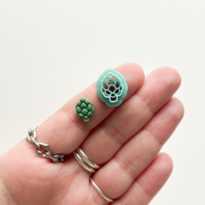 May include: Two green polymer clay earrings shaped like hops. The larger earring is a cutout design, while the smaller earring is solid.