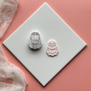 May include: Two white plastic cookie cutters shaped like the face of a monster. The cookie cutters are on a white surface.