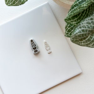 May include: Two lava lamp shaped cookie cutters, one is silver and the other is white. The cookie cutters are on a white tile surface.