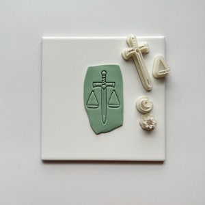 Può includere: Un set di cinque tagliapasta in plastica bianca, tra cui un design a spada e bilancia, una croce, un triangolo e due forme decorative. Il design a spada e bilancia è verde.