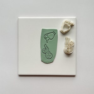 May include: A set of three white plastic cookie cutters, two are shaped like clouds and one is shaped like a pitcher pouring water. The cutters are on a white surface.