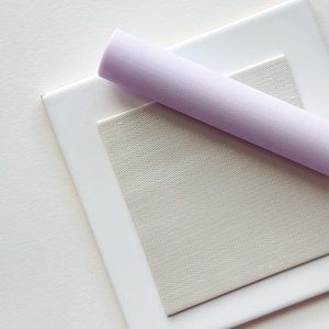 May include: A close-up of a white square frame with a light gray fabric inside. A light purple rolled fabric is placed on top of the frame.