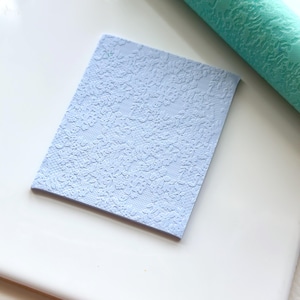 May include: A light blue square piece of polymer clay with a floral embossed pattern. The clay is sitting on a white surface.