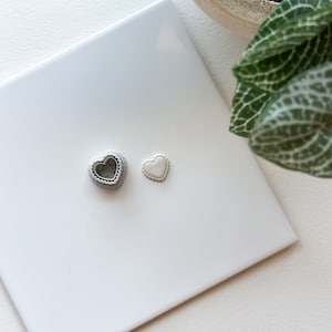 Puede incluir: Dos dijes de corazón plateados, uno con un centro hueco y el otro sólido, sobre un fondo blanco.
