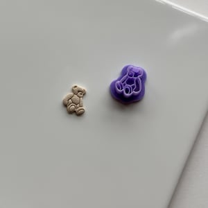 Pode incluir: Uma pequena figura de urso de pelúcia bege ao lado de um molde roxo em forma de urso. A figura do urso tem detalhes texturizados. O molde é de cor roxa sólida e tem a forma de um urso. Ambos os itens estão em uma superfície branca.