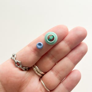 Puede incluir: Dos pequeños pendientes de arcilla polimérica, uno es un arándano azul claro y el otro es un cupcake verde y marrón con una flor roja en la parte superior.