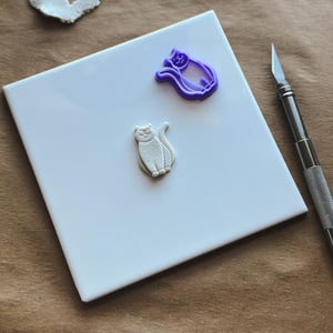 May include: A white ceramic tile with a white clay cat cutout and a purple plastic cat cookie cutter. A silver craft knife is on the right side of the image.