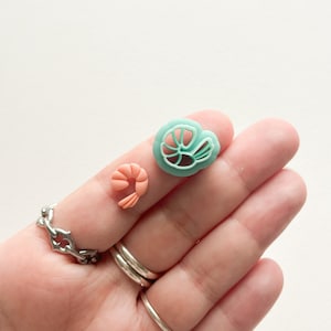 May include: Two polymer clay charms in the shape of shrimp. One charm is a light pink color and the other is a light teal color.