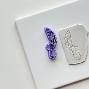 May include: Purple plastic cookie cutter shaped like a winged female torso. The cutter is on a white surface next to a clay impression of the cutter.