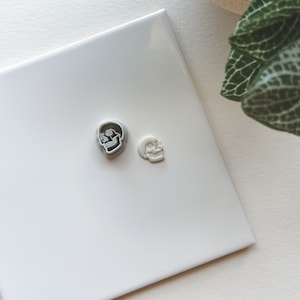 May include: Two small, silver-colored skull-shaped objects. One is larger and more detailed, while the other is smaller and simpler. They are placed on a white, glossy surface, possibly ceramic. The skulls are likely decorative or used in jewelry.