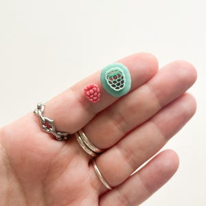 May include: Two polymer clay raspberry earrings, one is a solid pink color and the other is a light green color with a detailed raspberry design.