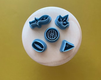 May include: Five blue plastic cookie cutters on a wooden surface. The cutters are shaped like a leaf, a circle with a design inside, a teardrop, a crescent moon, and a small circle.