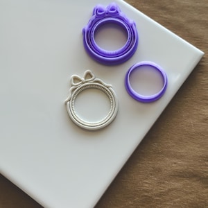May include: Three round cookie cutters, two are purple and one is white. The purple cutters have a bow design and the white cutter has a more ornate design.