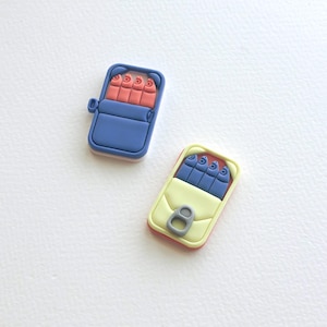 Op de afbeelding: Twee kleine, kleurrijke, plastic speelgoedblikjes sardines. Eén blikje is blauw met rode vissen en het andere is geel met blauwe vissen.