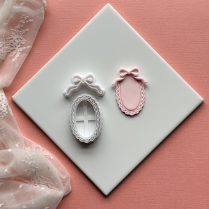 Op de afbeelding: Twee witte plastic koekjesvormen in de vorm van een ovaal met een strik bovenop. De vorm links heeft een kruisvormig ontwerp in het ovaal. De vorm rechts is massief.