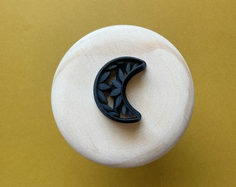 May include: A black crescent moon-shaped ornament with a decorative leaf pattern, displayed on a wooden surface.