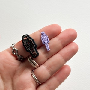 May include: Two robot-shaped charms in the image. One is black with red and silver details, the other is a light purple color. The charms are on a hand with silver rings. The rings have a chain-like design and a simple band.
