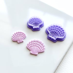 May include: Four purple and pink seashell-shaped cookie cutters. Two of the cutters are larger and have a detailed design, while the other two are smaller and have a more simple design.