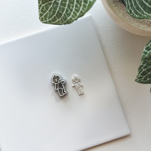 May include: Two small ceramic figurines of babies, one in white and one in grey, on a white surface.
