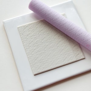 May include: A white square tile with a raised brick pattern texture. A purple textured rolling pin is laying on top of the tile.