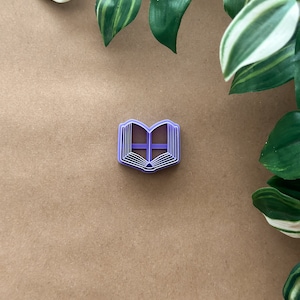 May include: Purple plastic cookie cutter shaped like an open book. The cookie cutter is on a brown surface.