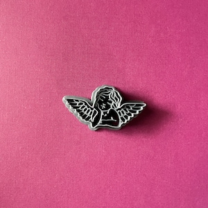 May include: A black and white angel pin with wings outstretched. The angel is depicted with a simple, stylized design.