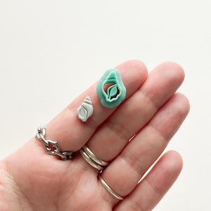 Op de afbeelding: Een hand houdt twee schelpvormige polymeerklei-uitsteekvormen en een kleine schelp vast. De uitsteekvormen zijn teal en de schelp is wit en grijs. De hand draagt ook verschillende zilveren ringen en een kettingarmband.