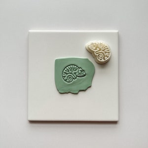 Könnte beinhalten: Ein weißer Keramik-Chamäleon-Stempel und ein grüner Tonabdruck des Stempels. Das Chamäleon ist zusammengerollt, mit dem Schwanz um den Körper gewickelt.