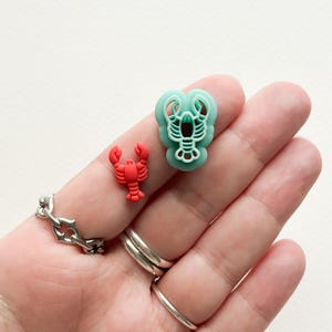 May include: Two lobster-shaped charms, one red and one teal, are held in a hand. The charms are small and detailed.