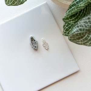 May include: Two silver charms in the shape of a hand with fingers curled inward. The charms are on a white tile surface.