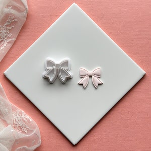 Puede incluir: Dos cortadores de galletas de plástico blanco en forma de lazos. El lazo más pequeño es rosa.