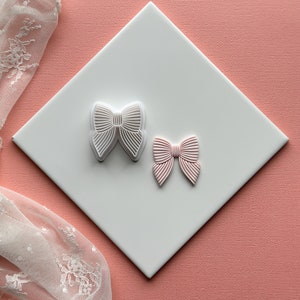 Puede incluir: Dos cortadores de galletas de plástico blanco en forma de lazo, uno con un diseño en relieve y otro con una superficie lisa. El cortador con el diseño en relieve está sobre una superficie blanca, y el cortador liso está sobre una superficie rosa.