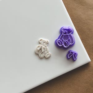 Op de afbeelding: Twee paarse plastic beren koekjesvormen en een witte plastic beren koekjesvorm op een wit oppervlak.