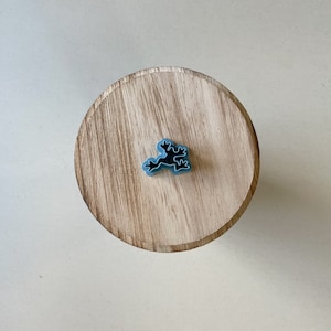 Może przedstawiać: Jasnoniebieski, stylizowany charms w kształcie żaby na okrągłej, drewnianej powierzchni. Charms jest wyśrodkowany na drewnie, które ma naturalny wzór słojów. Tło jest jednolite, w kolorze złamanej bieli.