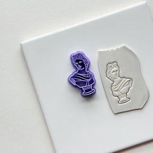 May include: A purple plastic stamp of a bust of a man with a laurel wreath on his head. The stamp is on a white surface next to a white clay impression of the stamp.