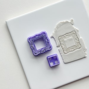 Pode incluir: Três cortadores de biscoitos de plástico roxo em forma de molduras. A moldura maior tem um design decorativo. A moldura menor é um quadrado simples com um padrão de grade.