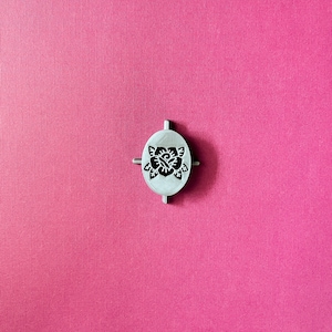 May include: A silver metal cookie cutter in the shape of a flower with four arms. The cookie cutter is on a pink background.