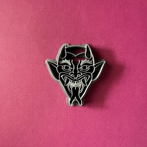 May include: A black and white cookie cutter in the shape of a demon's face with horns, fangs, and a tongue sticking out.