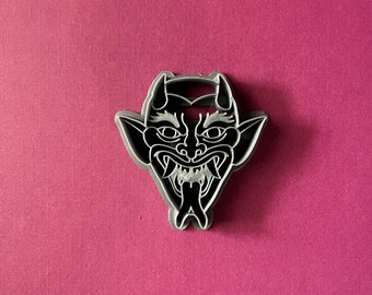 May include: A black and white cookie cutter in the shape of a demon's face with horns, fangs, and a tongue sticking out.