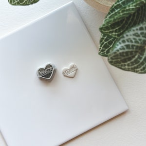May include: Two small heart-shaped charms with the words "Juno" and "Amari" on them. The charms are on a white surface.