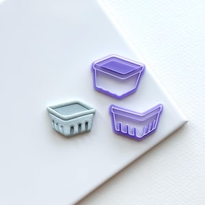 May include: Three miniature basket-shaped objects in the image. One is a light gray color with a textured surface, while the other two are purple with a transparent appearance. The objects are arranged on a white surface.