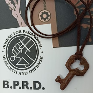 May include: A brown key pendant on a brown cord. The key has a rustic, aged appearance. The background includes a logo with the text "BUREAU FOR PARANORMAL RESEARCH AND DEFENSE" and the letters "B.P.R.D."