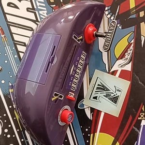 Puede incluir: Una consola de juegos Zathura morada con botones rojos y un panel deslizante. La consola tiene una pantalla digital y una manivela. El fondo presenta una ilustración de temática espacial con la palabra "Zathura" y un cohete.