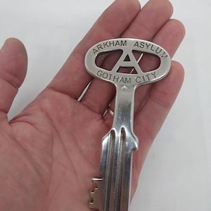 May include: A silver key featuring the words "ARKHAM ASYLUM" and "GOTHAM CITY". The key has an "A" design and a detailed key bit. The key is held in a hand.