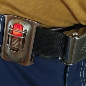 May include: A black belt with a silver buckle and a silver and brown device with a red light. The device is attached to the belt.